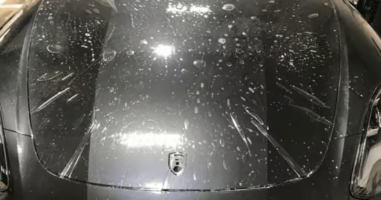 Black sports car hood and windshield covered in water droplets under bright lights