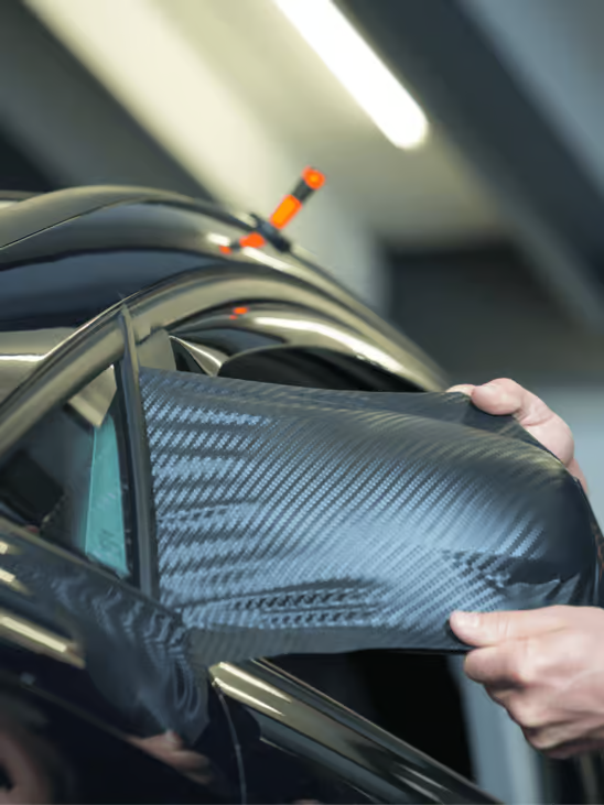 Hands fitting a carbon-fiber side mirror cover on a black car indoors