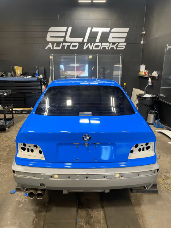Blue BMW race car in Elite Auto Works shop, rear view with exposed taillights and bumper removed