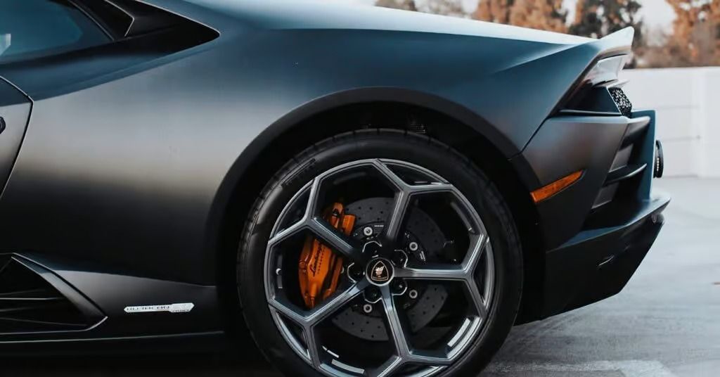 Close-up of a matte black sports car front wheel and fender, with orange brake caliper on a driveway