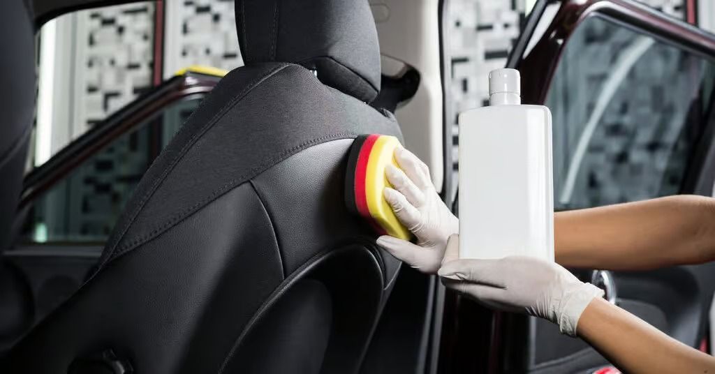 Gloved hands cleaning a car seat with a sponge and spray bottle inside a vehicle