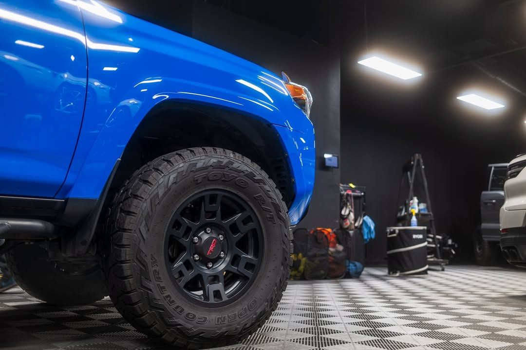 Blue lifted truck in a garage with off-road tires and a checkered floor, with another vehicle in the background