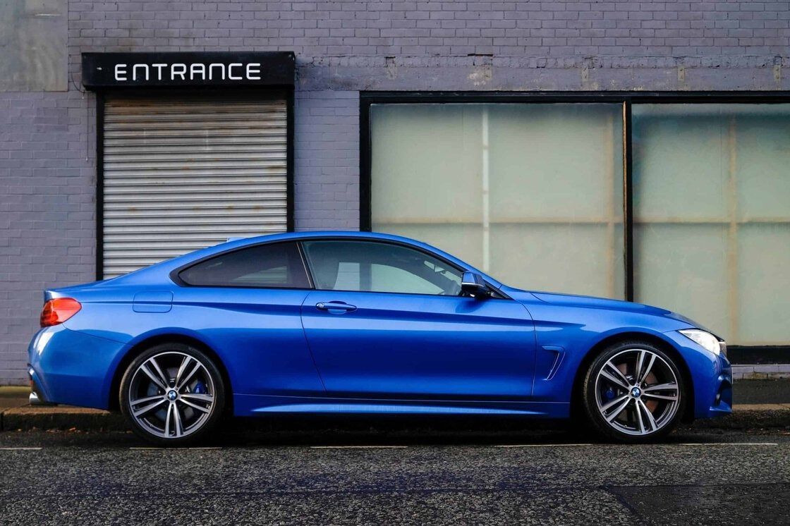 Blue sports coupe parked beside a gray building with a closed entrance and large windows.