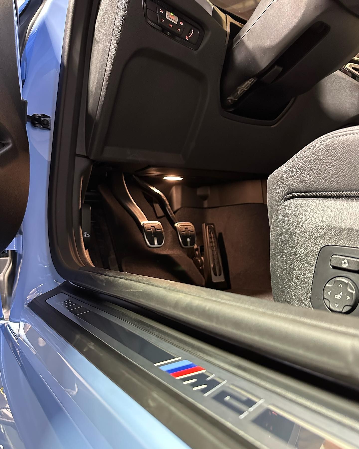 Open car door showing a black and brown interior with M badge on the sill and seat controls visible