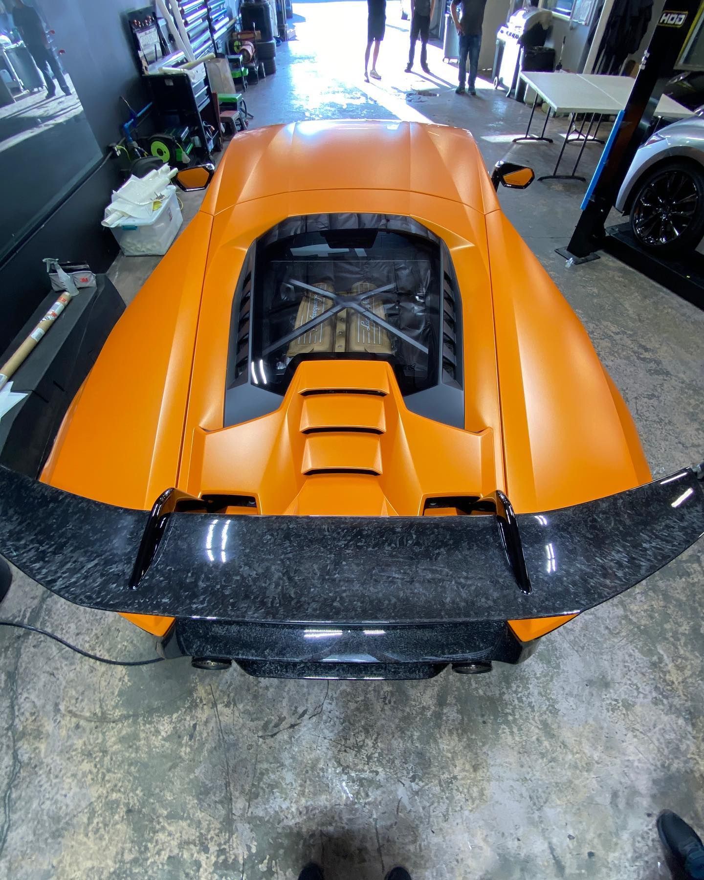 Orange sports car with exposed rear engine and large black rear wing in a garage