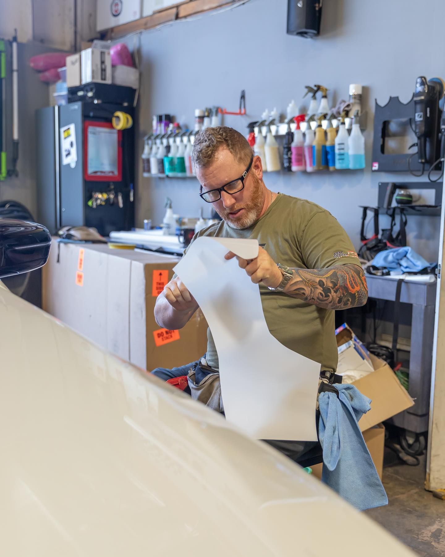Tattooed worker examining a white sheet in a workshop with paint bottles and tools on shelves