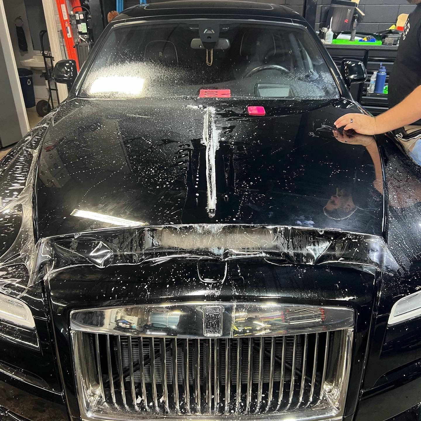Black car being washed in a garage, with water and soap on the hood and a hand reaching in from the right.