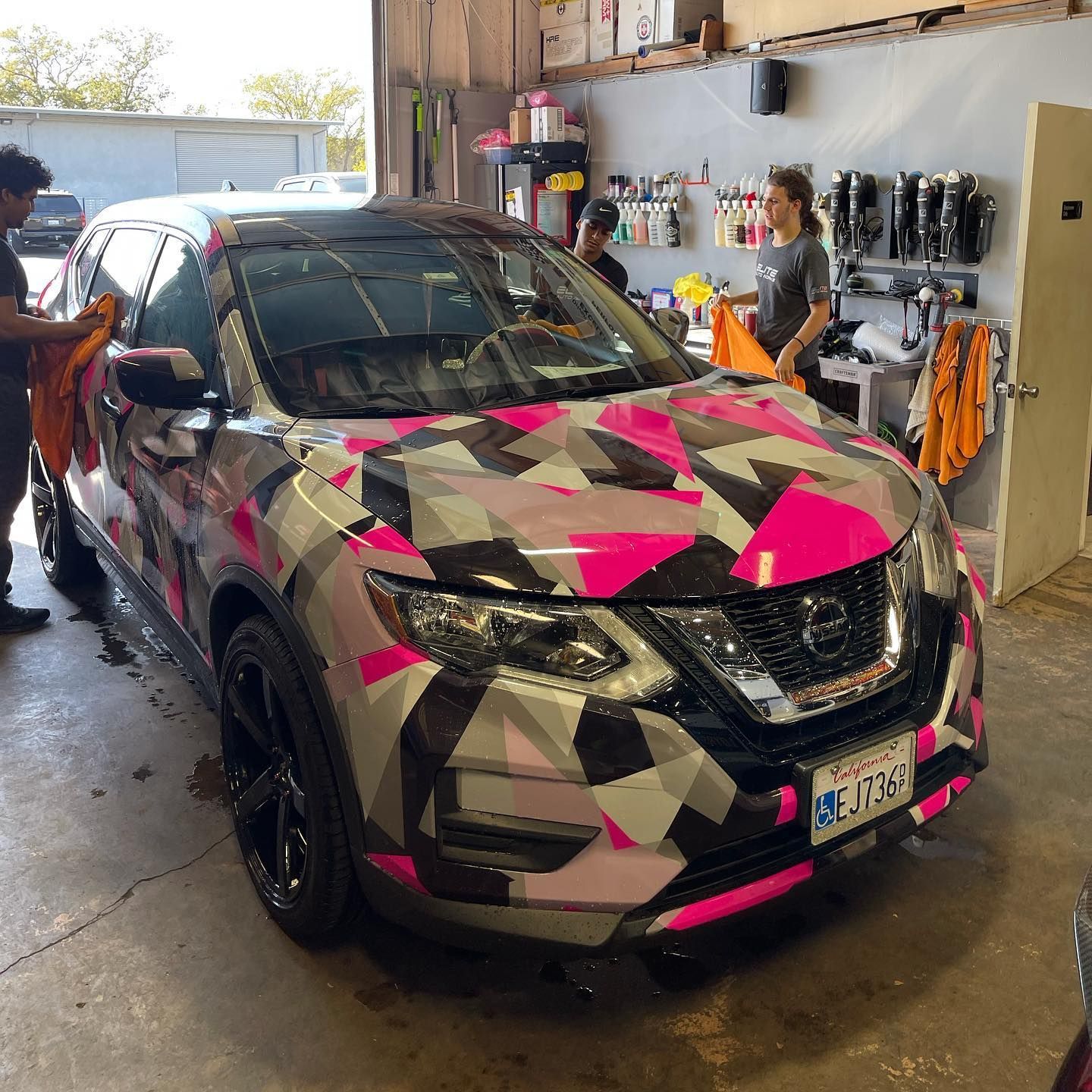 Camouflaged Nissan SUV with pink, gray, and black wrap in an auto shop, with two people nearby
