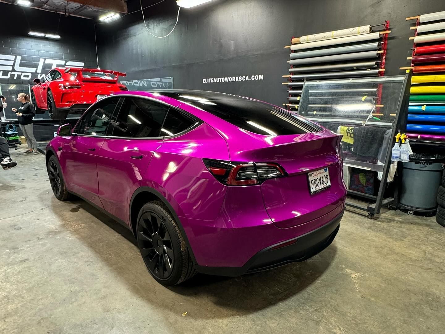 Purple Tesla SUV in an auto shop, parked under bright lights with colorful vinyl rolls on the wall.