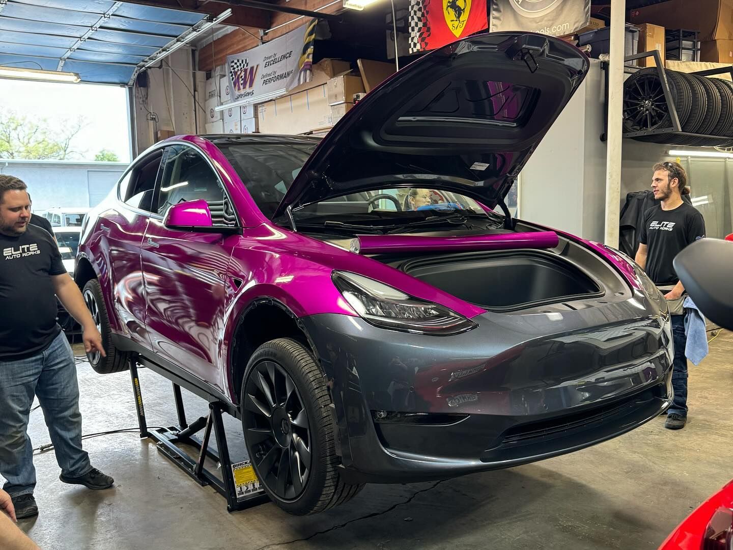 Magenta car on a lift in a garage with its hood open, two people standing nearby