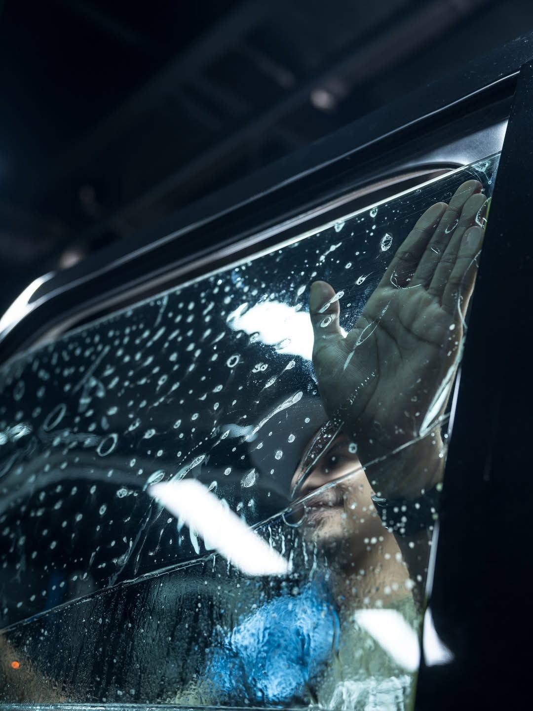Hand against rain-speckled car window at night, with bright reflections inside the vehicle