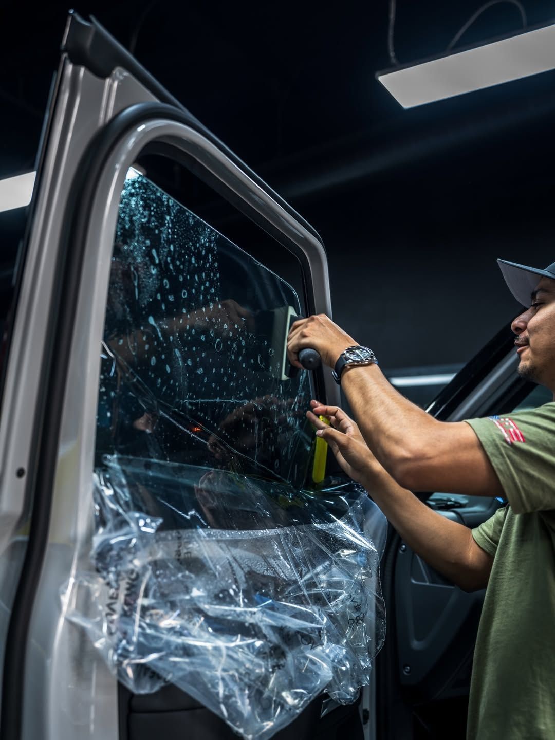 Technician working on a car window, applying film or repair material in a garage.