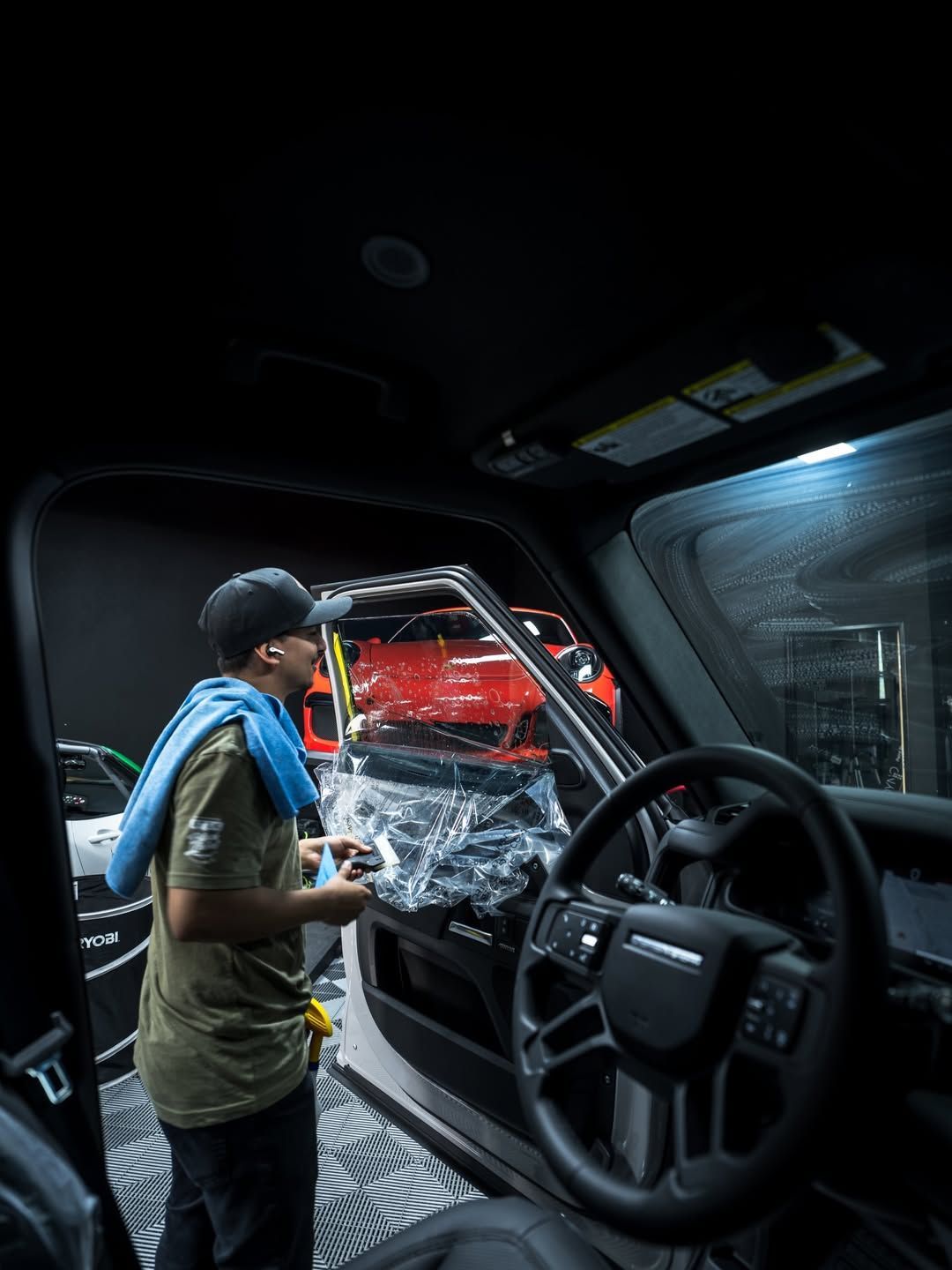 Person leaning into a vehicle, holding a tool near a shattered windshield, with red light outside.