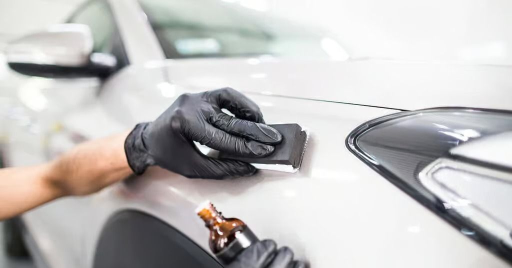 Black-gloved person sanding a silver car body panel in an auto shop