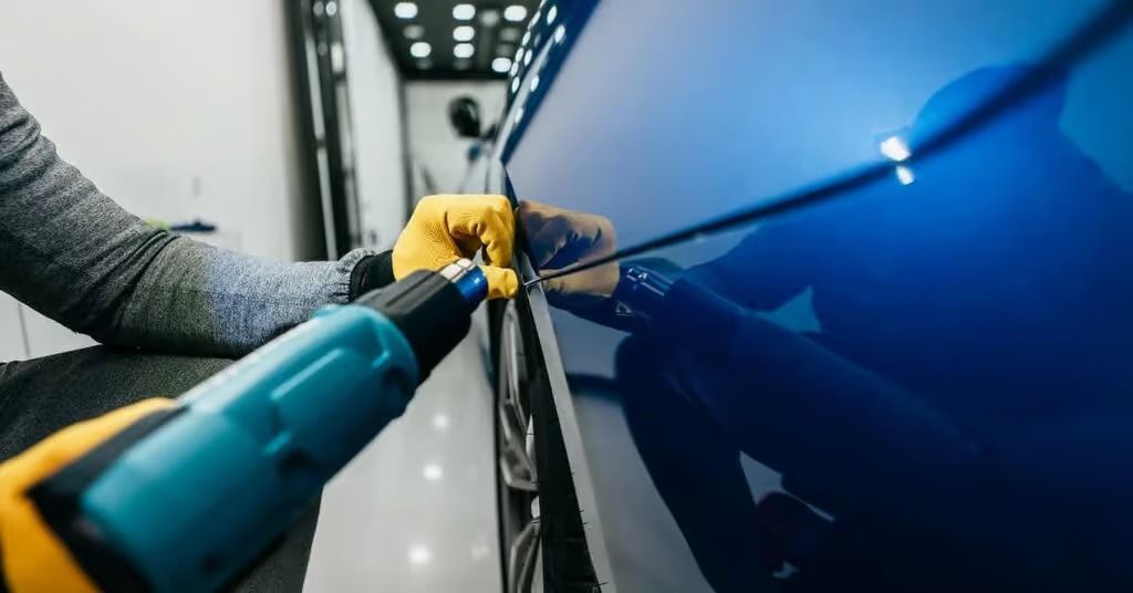Person polishing a blue car door with a buffer, wearing yellow gloves in a workshop