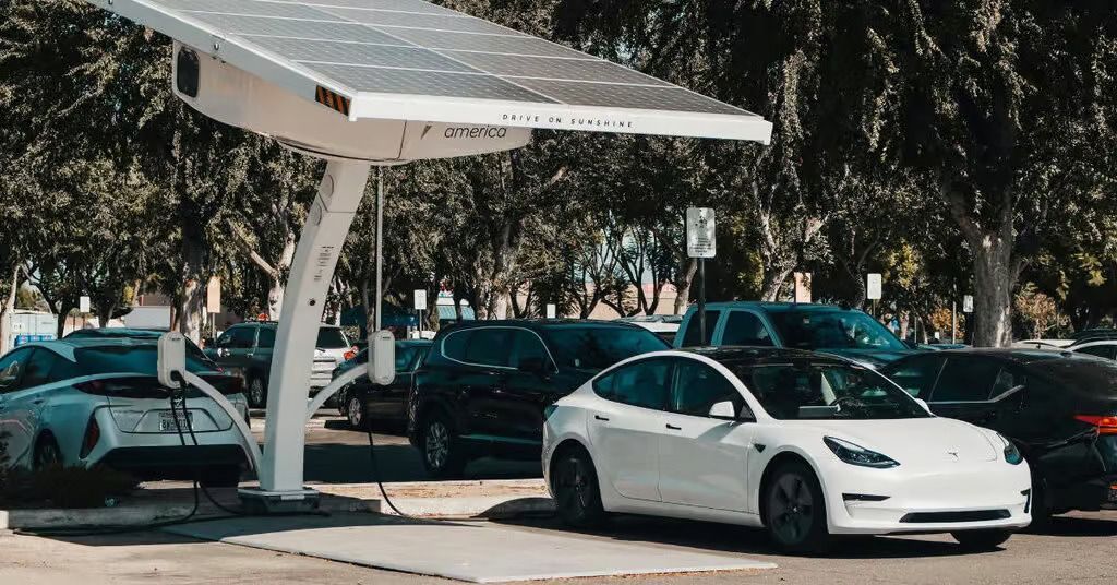White sedan parked under a solar canopy in a lot, with several cars and trees in the background