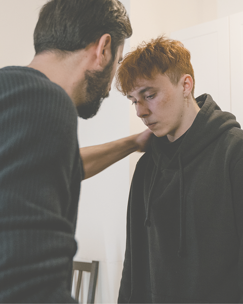 Man comforting a younger man with red hair; setting is indoors.