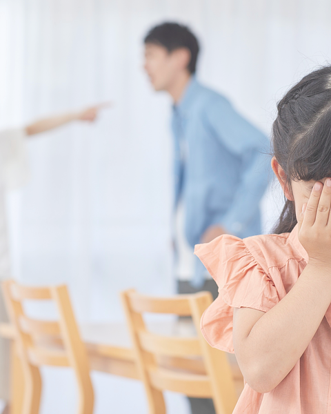A young girl covers her ears while parents argue in the background.