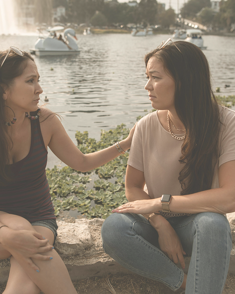 Two women by a lake, one comforting the other. White swan boats in background.