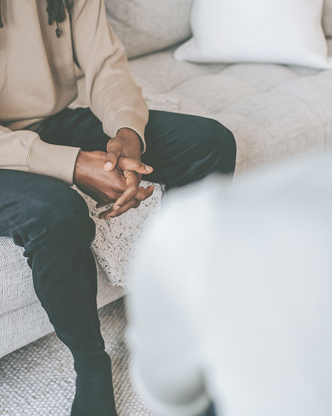 Person sitting on a couch, hands clasped, interacting with another person out of focus. Beige and black attire.
