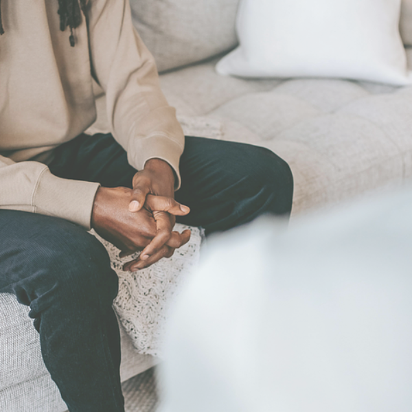 Person sitting on a gray couch with hands clasped. They wear black pants and a tan sweater. Soft lighting.