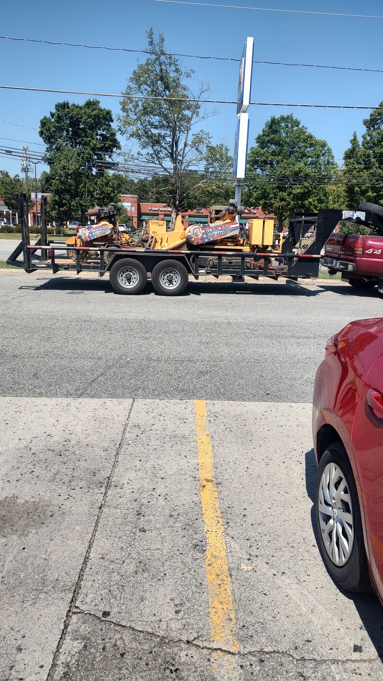 Yellow construction equipment on a flatbed trailer parked along a street, with trees and cars nearby