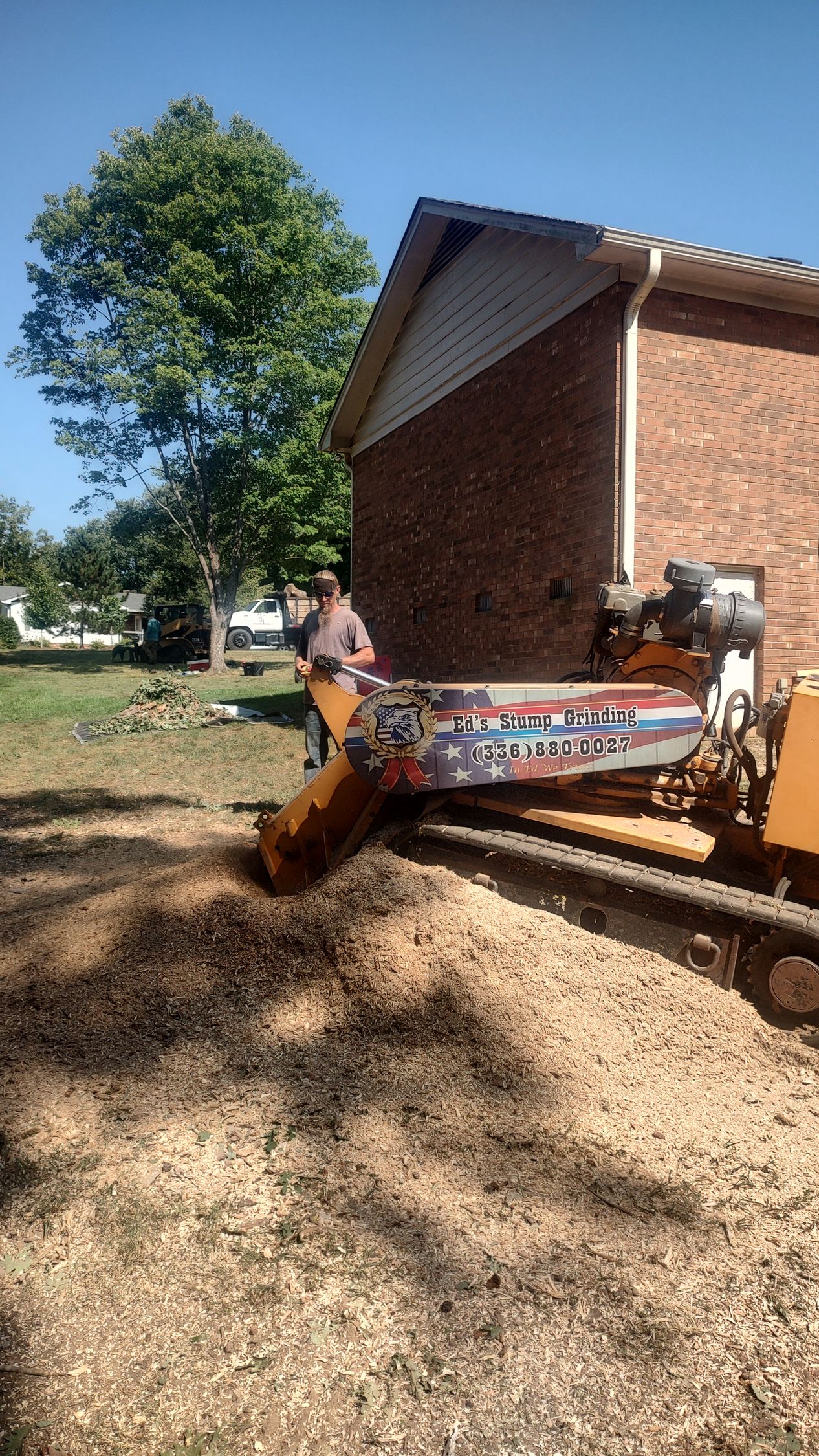 Small excavator clearing dirt beside a brick building under a clear blue sky.
