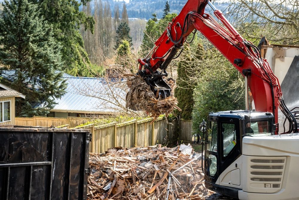 An excavator with a grapple arm, lifting debris from a demolition site into a dumpster.