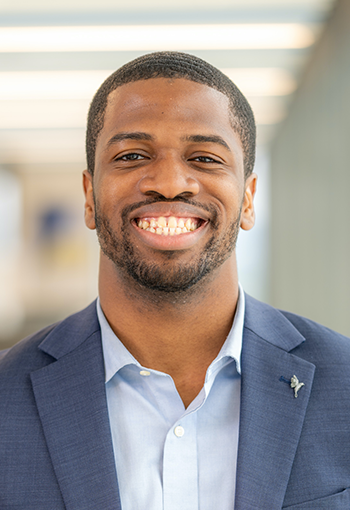 Smiling man in a blue blazer and light blue shirt standing indoors in a bright hallway