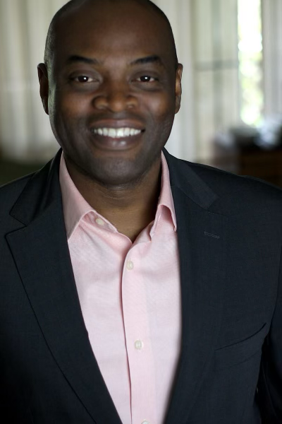 Smiling person in a dark blazer and light pink shirt, standing indoors with a blurred background