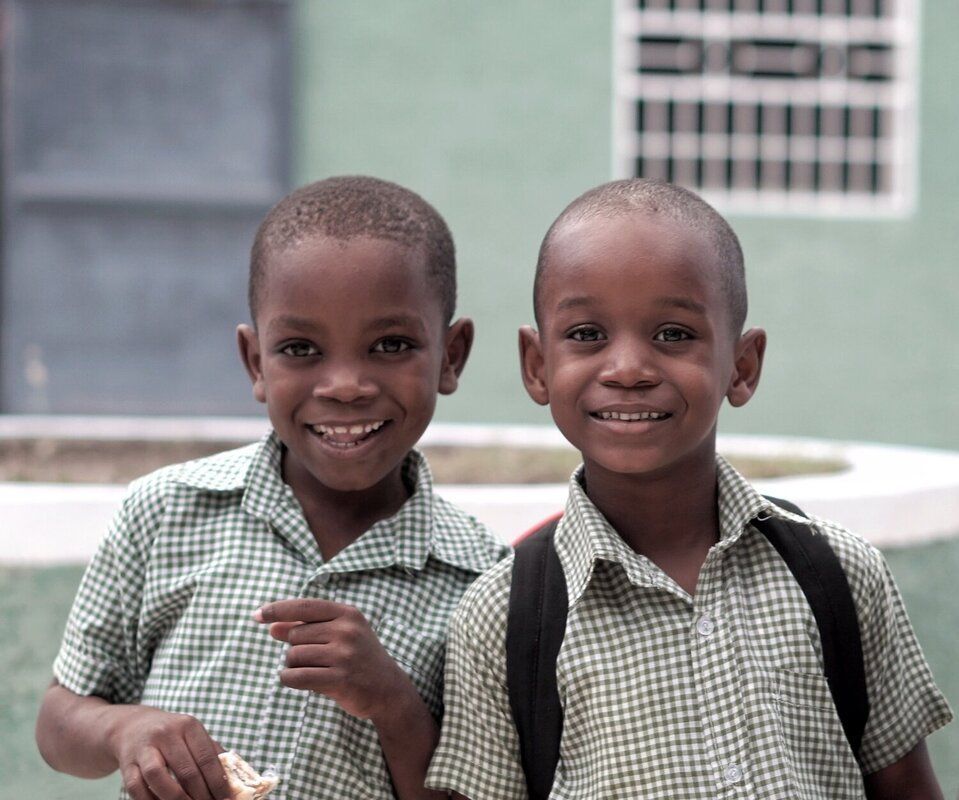 Two schoolchildren in green checkered uniforms smiling outdoors, one carrying a backpack