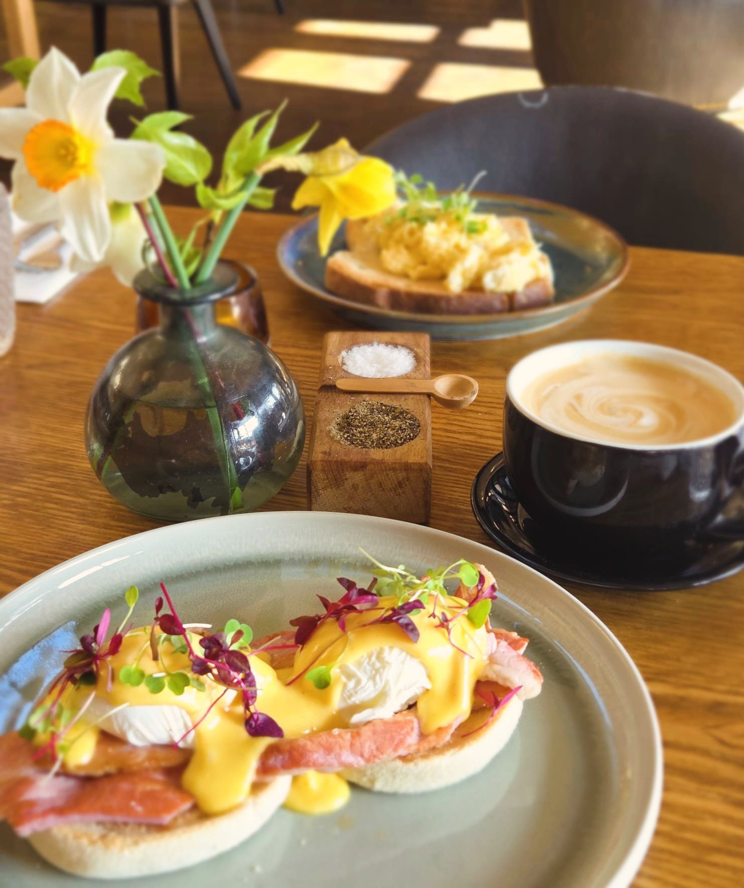 Brunch spread with eggs benedict, avocado toast, latte, and flowers on a sunlit table
