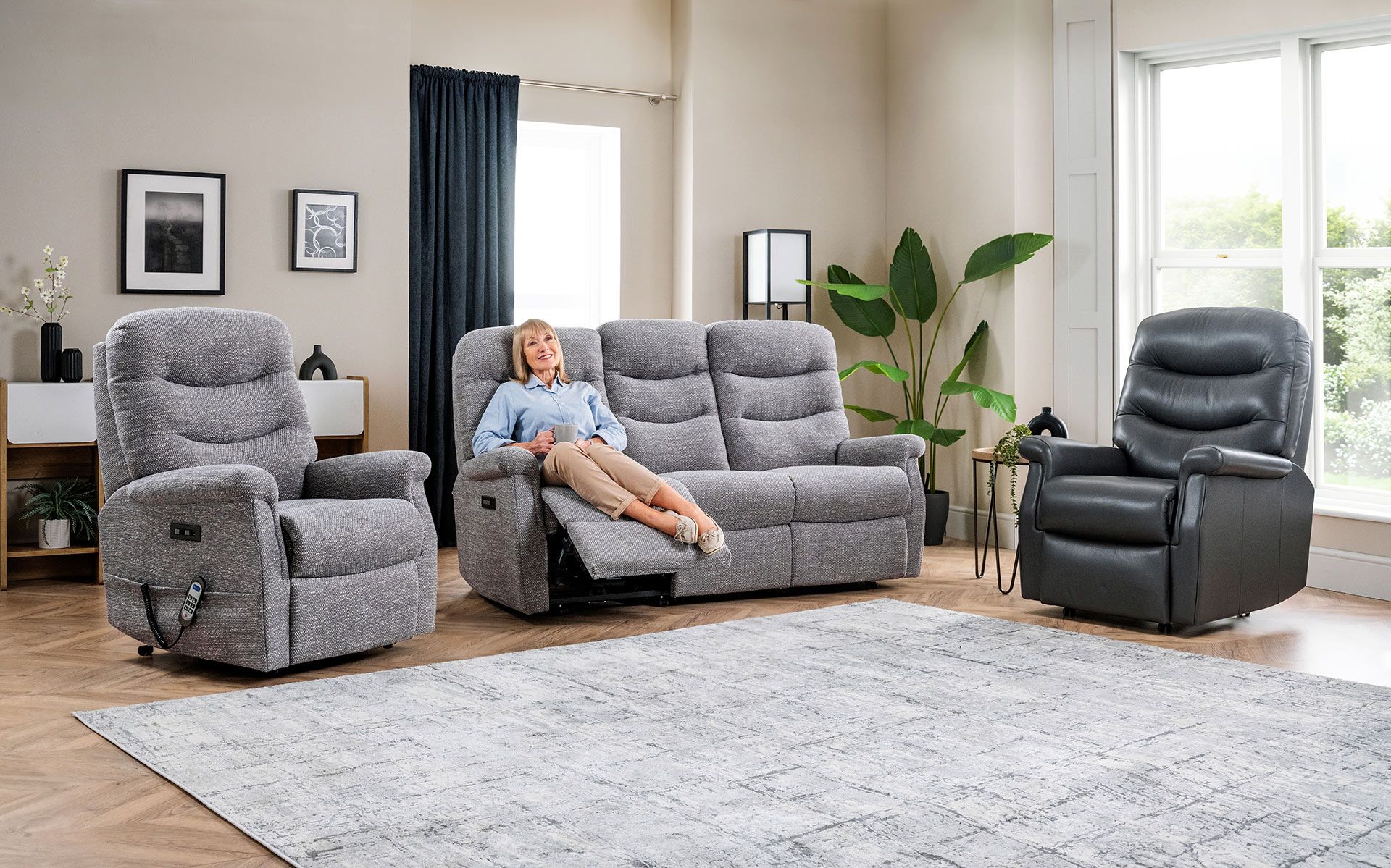 Living room with three gray recliners, a person lounging on a sofa, and a large rug by a bright window