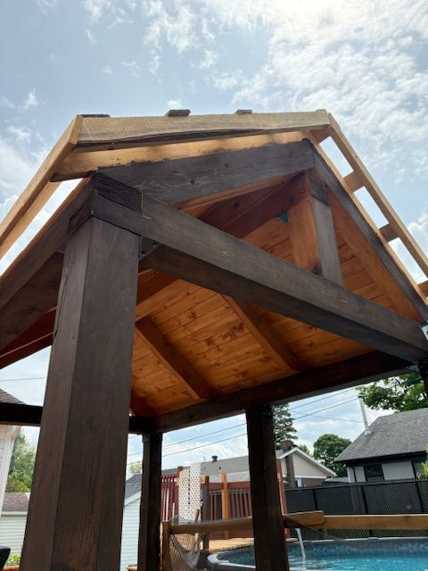 Pergola en bois en construction, teintée en brun, avec une charpente de toit en bois naturel se détachant sur un ciel bleu.