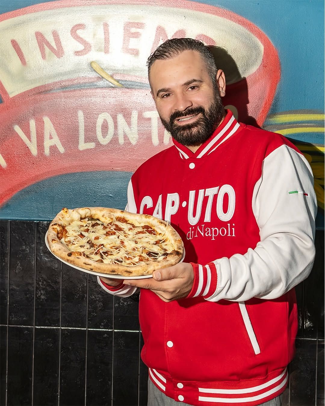 Uomo con una giacca rossa che tiene una pizza davanti a un murale di un ristorante