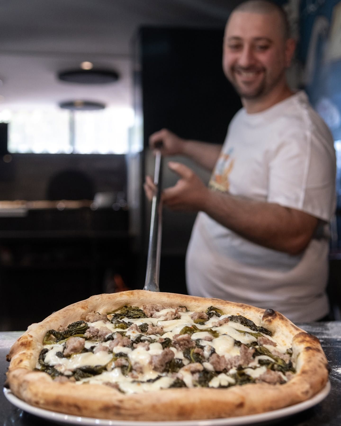 Uno chef sorride mentre prepara una pizza in cucina, con una pizza appena sfornata in primo piano.