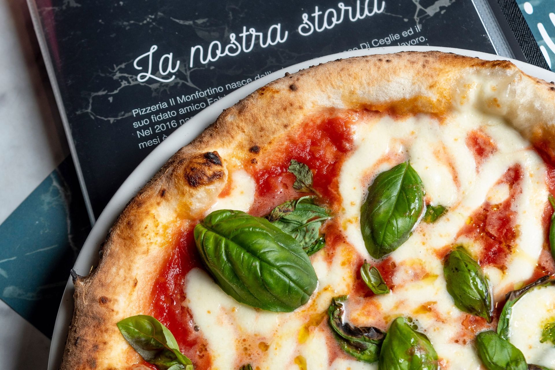 Pizza margherita con basilico su un piatto accanto a una lavagna con la scritta «La nostra storia»