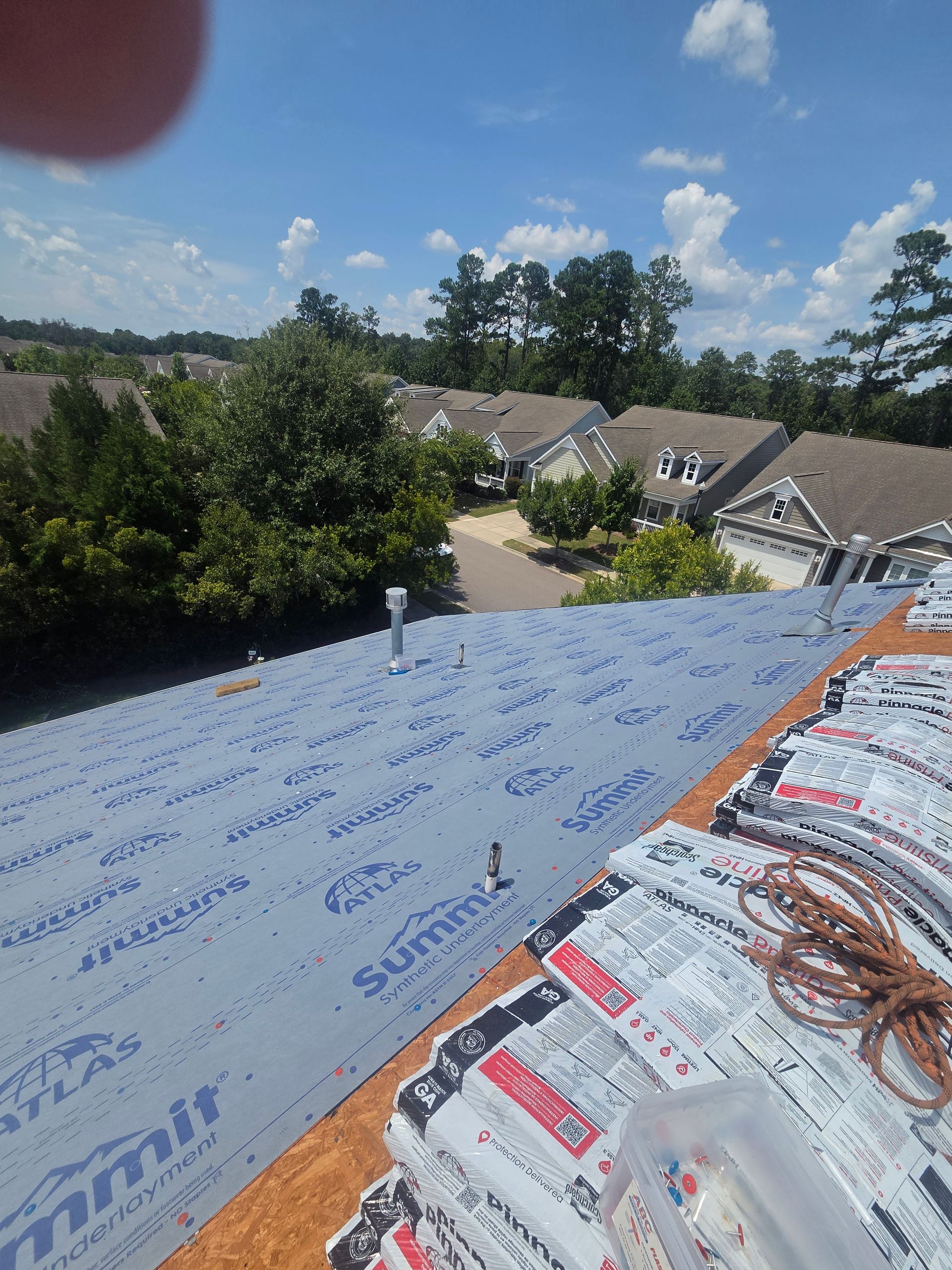 A Charleston Roofs and Windows crew installing a new standing seam metal roof on a one-story ranch home in Ravenel, SC, set back from the road on a wooded lot with tall loblolly pines and a spreading live oak in the yard. A W2 project manager walks the gutter line with the homeowner, representing the roofing company serving Ravenel SC for roof inspections, roof repairs, roof replacements, siding, windows and gutters, rated 4.9 stars on 680 Google reviews.