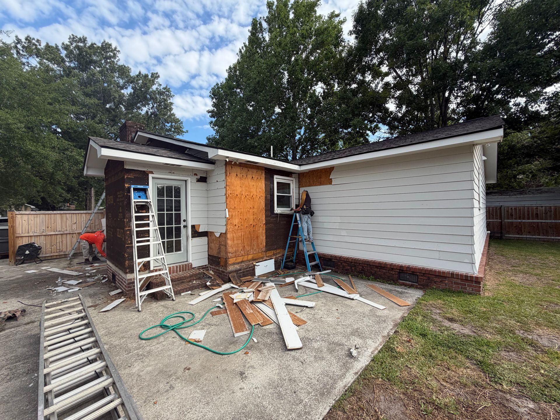 A Charleston Roofs and Windows inspection manager reviewing a line-item roof replacement estimate with a Ladson, SC couple on the front porch of a single-story brick ranch home, with a new architectural shingle roof overhead and pine trees in the yard. The image represents roof replacement cost in Ladson SC, priced per square for asphalt shingles, metal and flat roofs, with free inspections and 0% financing for 12 months on approved credit.