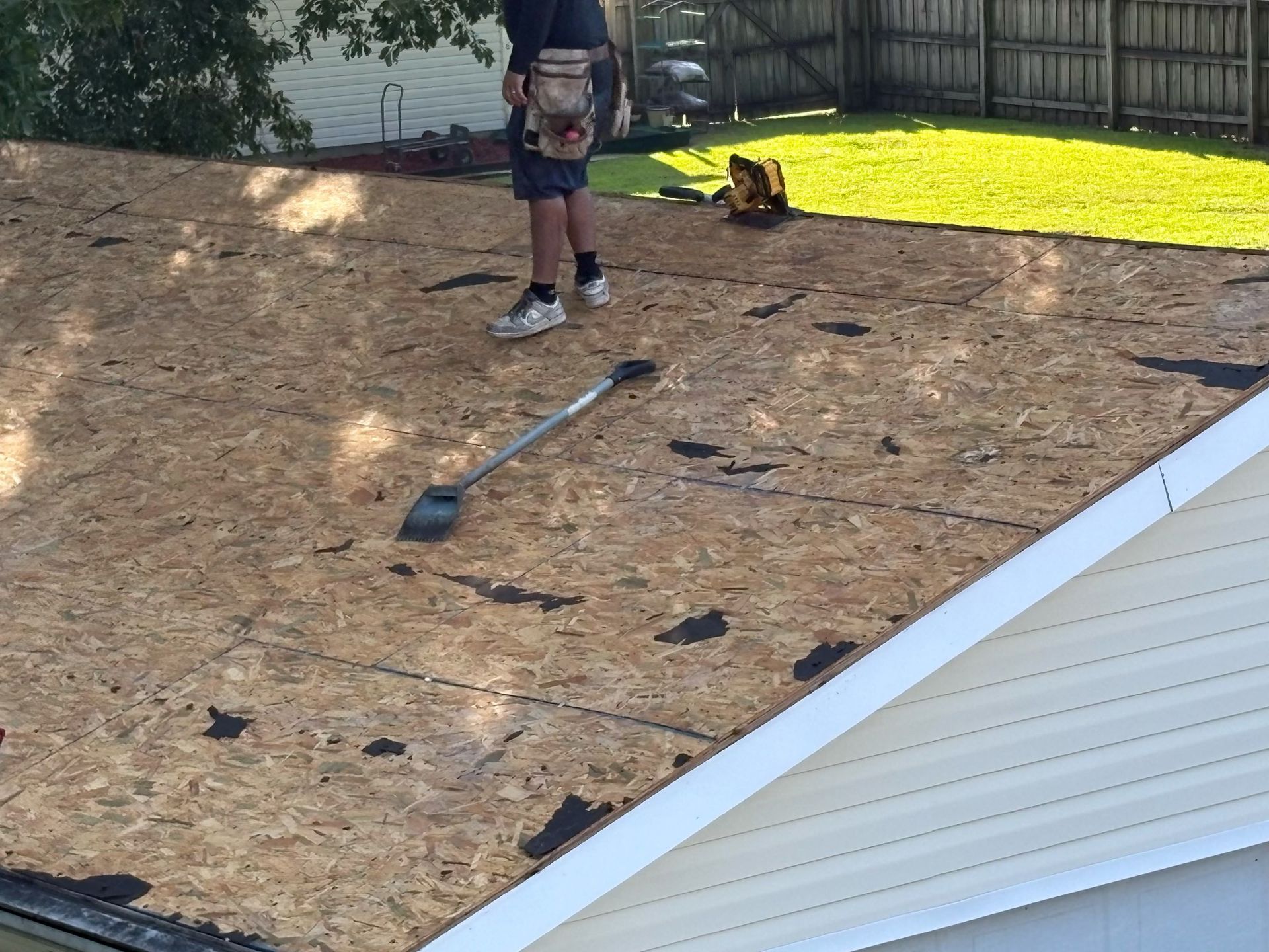 A Charleston Roofs and Windows inspection manager on a ladder photographing ridge cap and pipe boot flashing on a one-story brick ranch home in Hollywood, SC, with tall loblolly pines behind the house. The image shows the free roof inspection that starts every everyday roof repair and storm damage project in Hollywood, SC, from a locally owned roofing company working in the Lowcountry since January 2012.