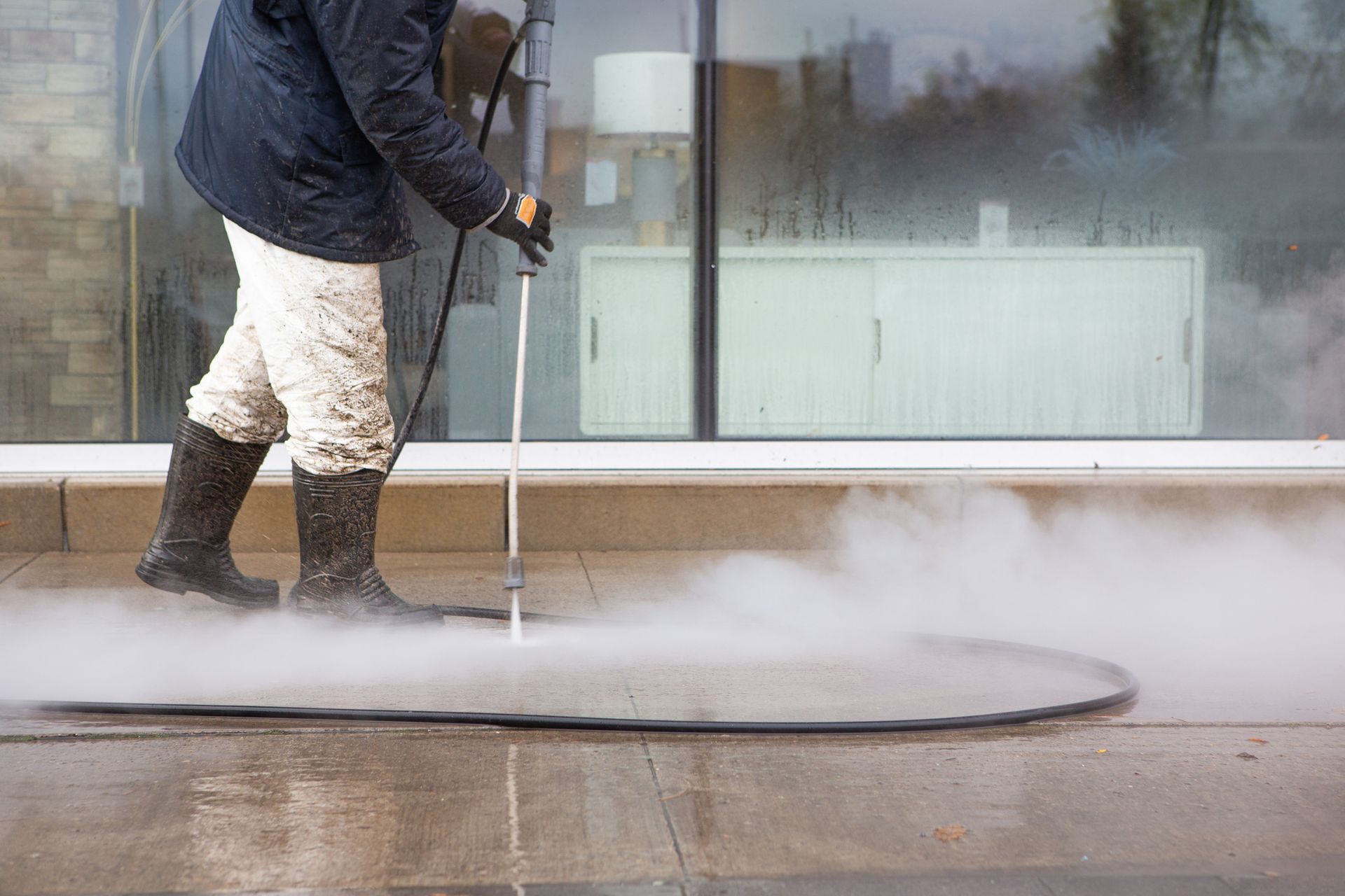 Worker power washing the sidewalk wearing black rubber boots and white pants.