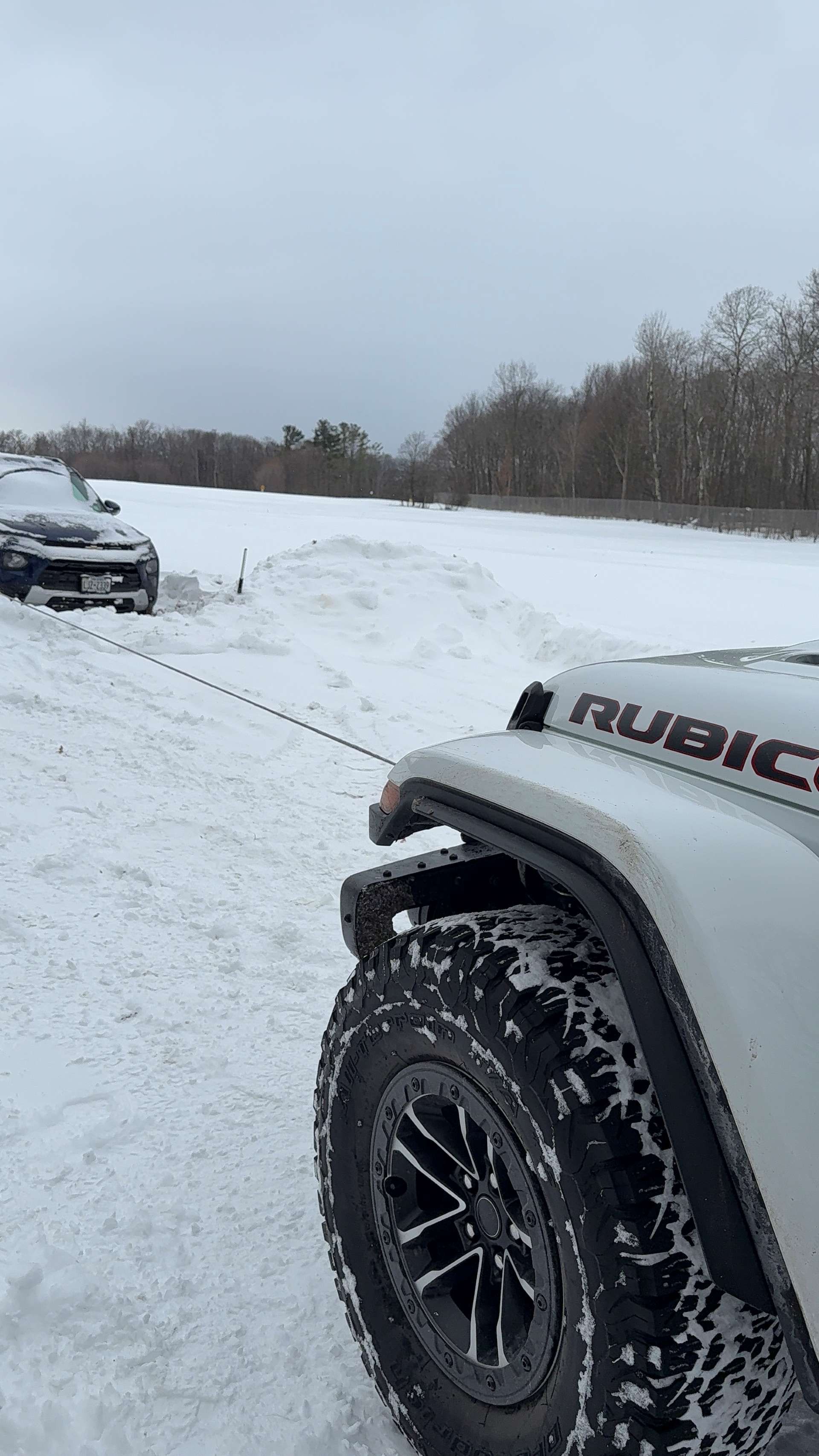A white Jeep Rubicon winching a vehicle stuck in deep snow on a bleak, overcast day.