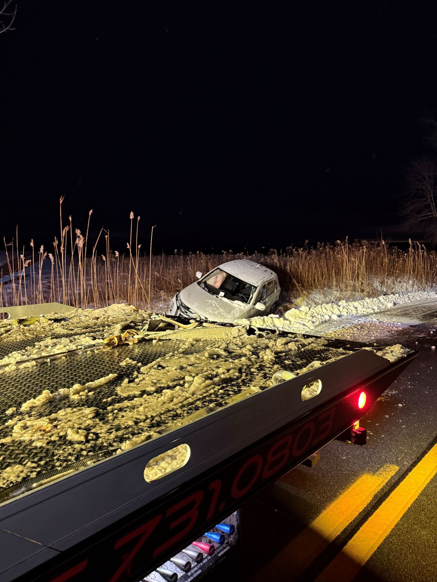 A silver car sits in a snow-covered ditch at night, viewed from the rear of a parked tow truck.