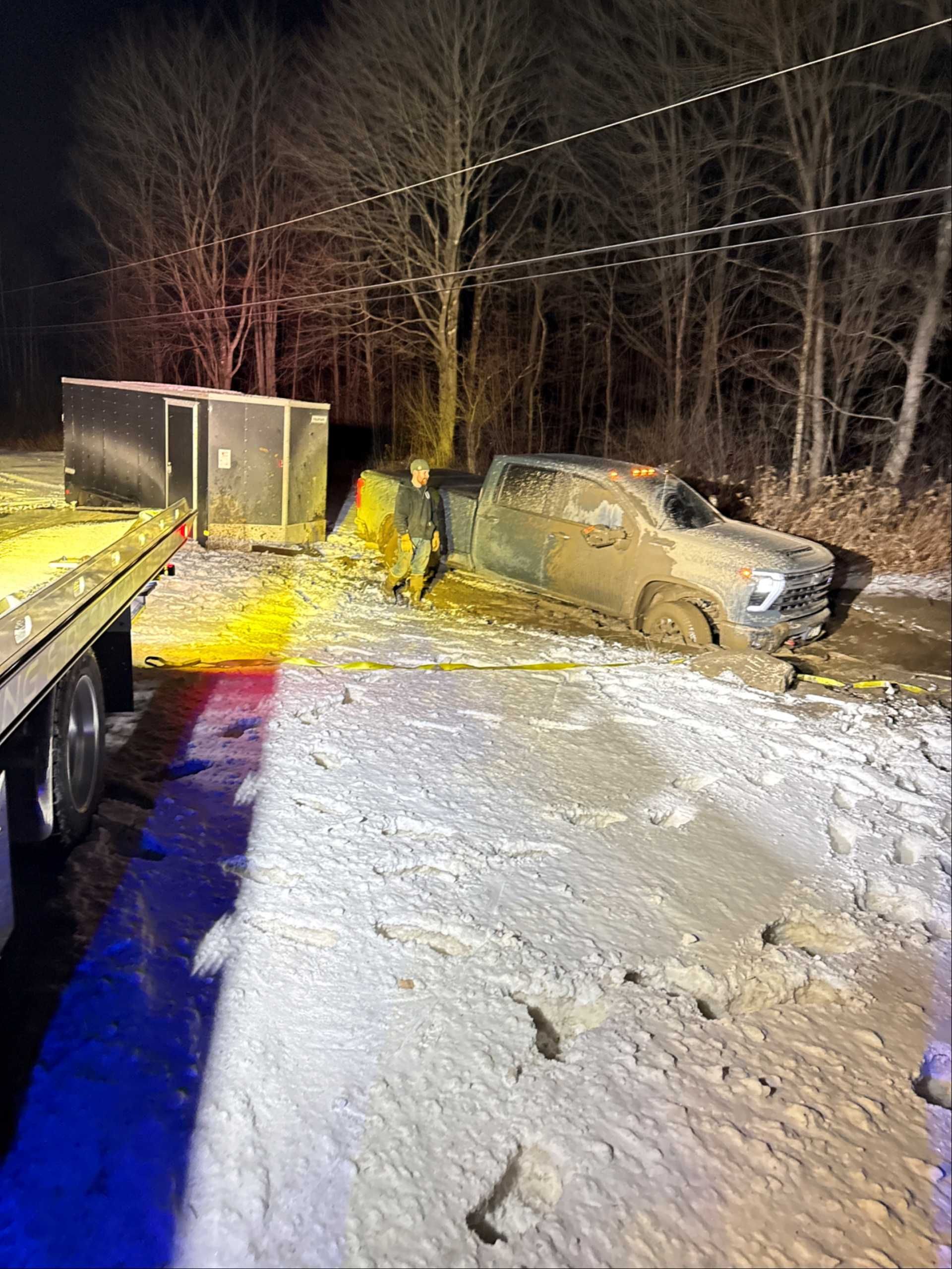 A truck and a jackknifed trailer are stuck in mud and snow on a dark roadside next to an emergency vehicle.