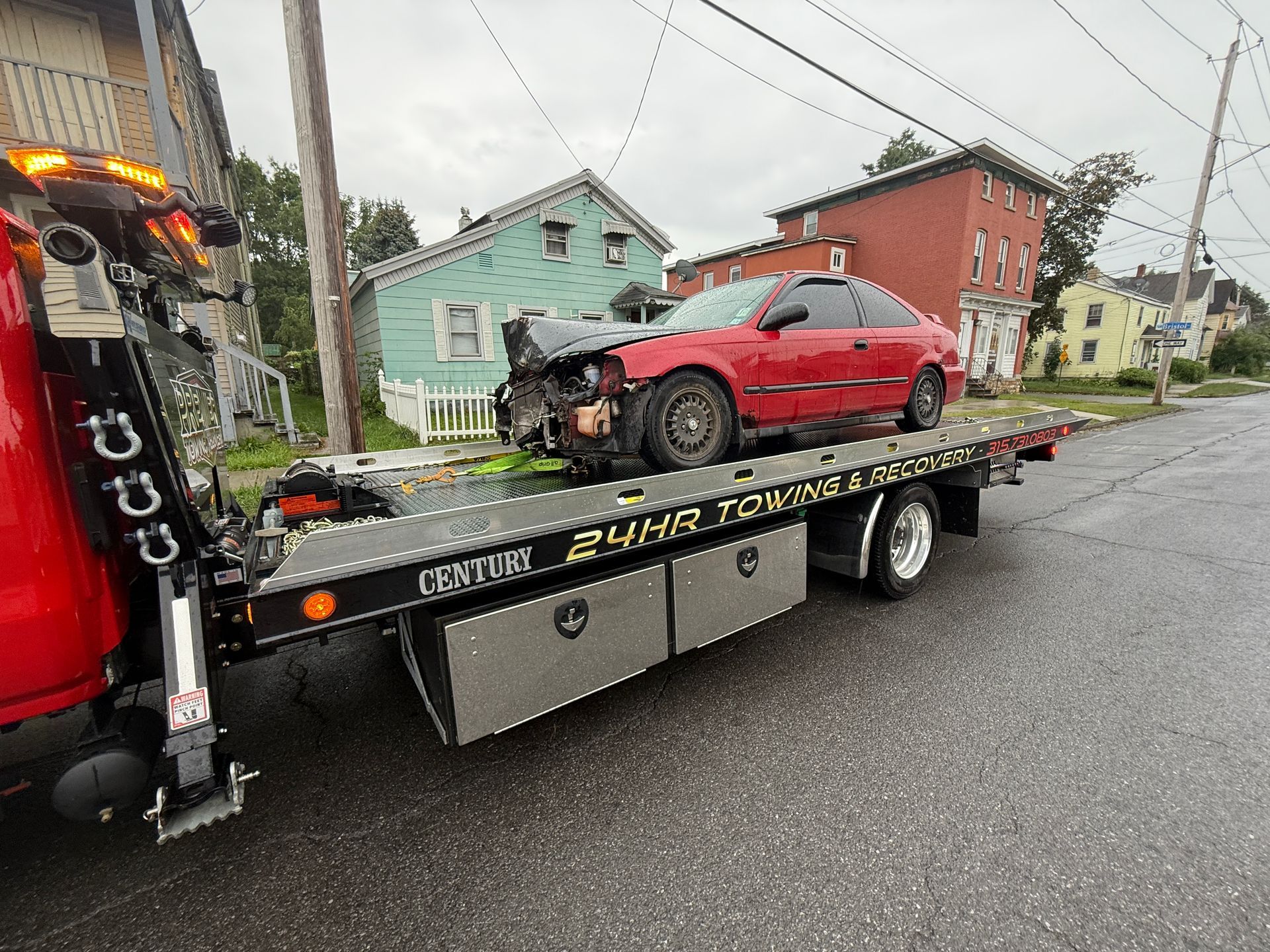A badly damaged red car is secured on a flatbed tow truck on a residential street.