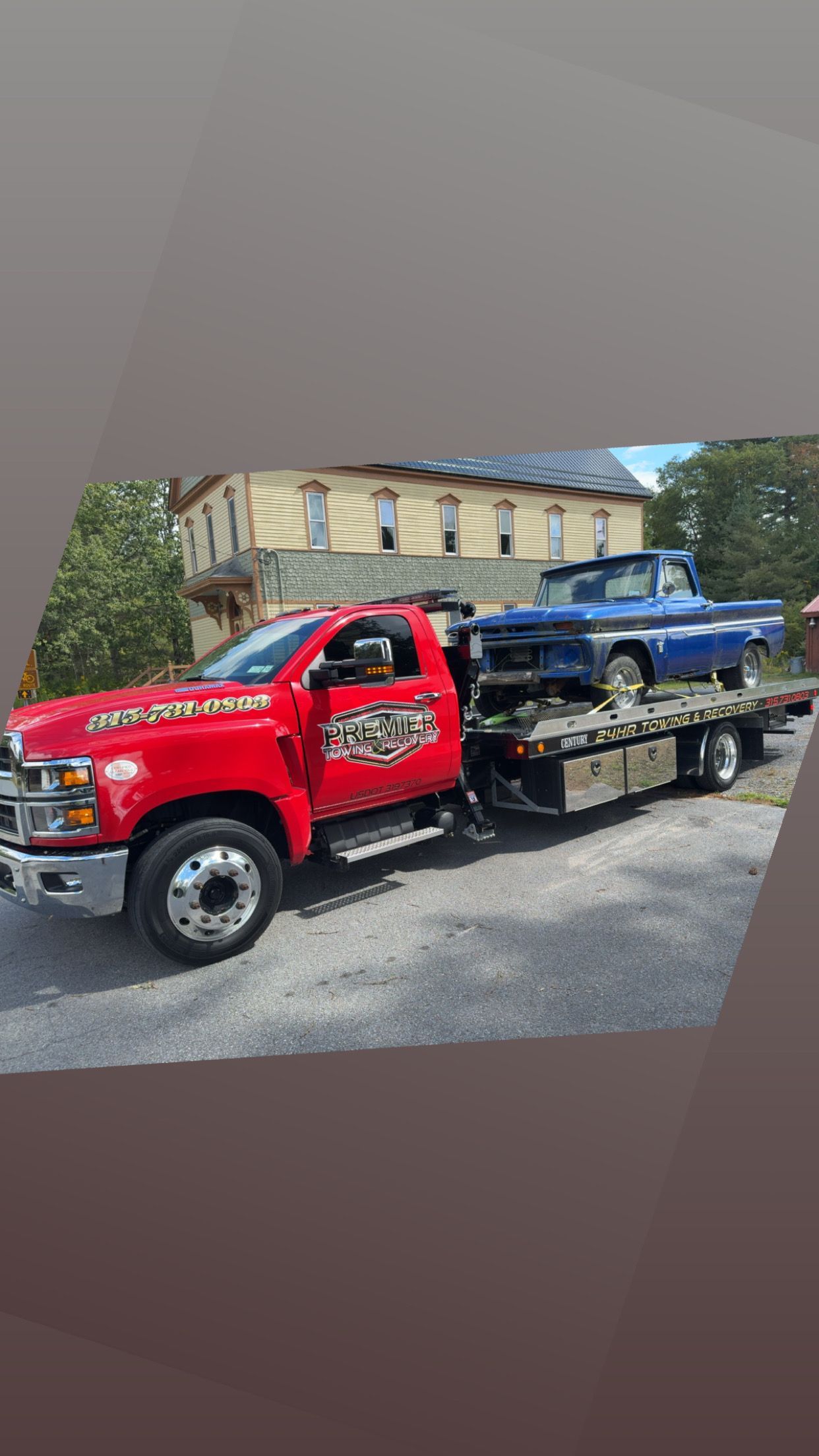 A bright red flatbed tow truck transporting a vintage blue pickup truck in front of a building.