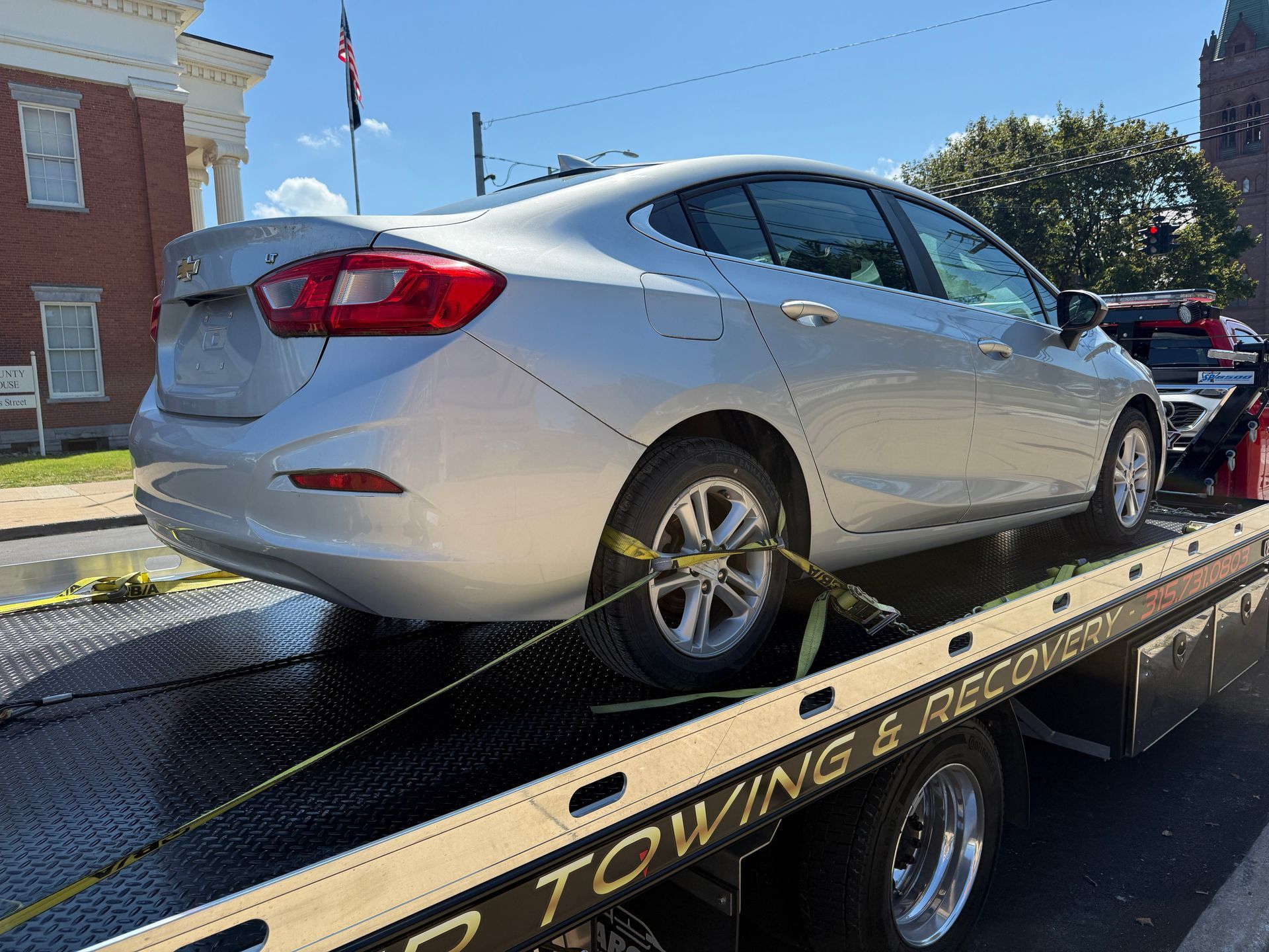 A silver sedan is strapped down and secured on the flatbed of a tow truck on a sunny day near a brick building.