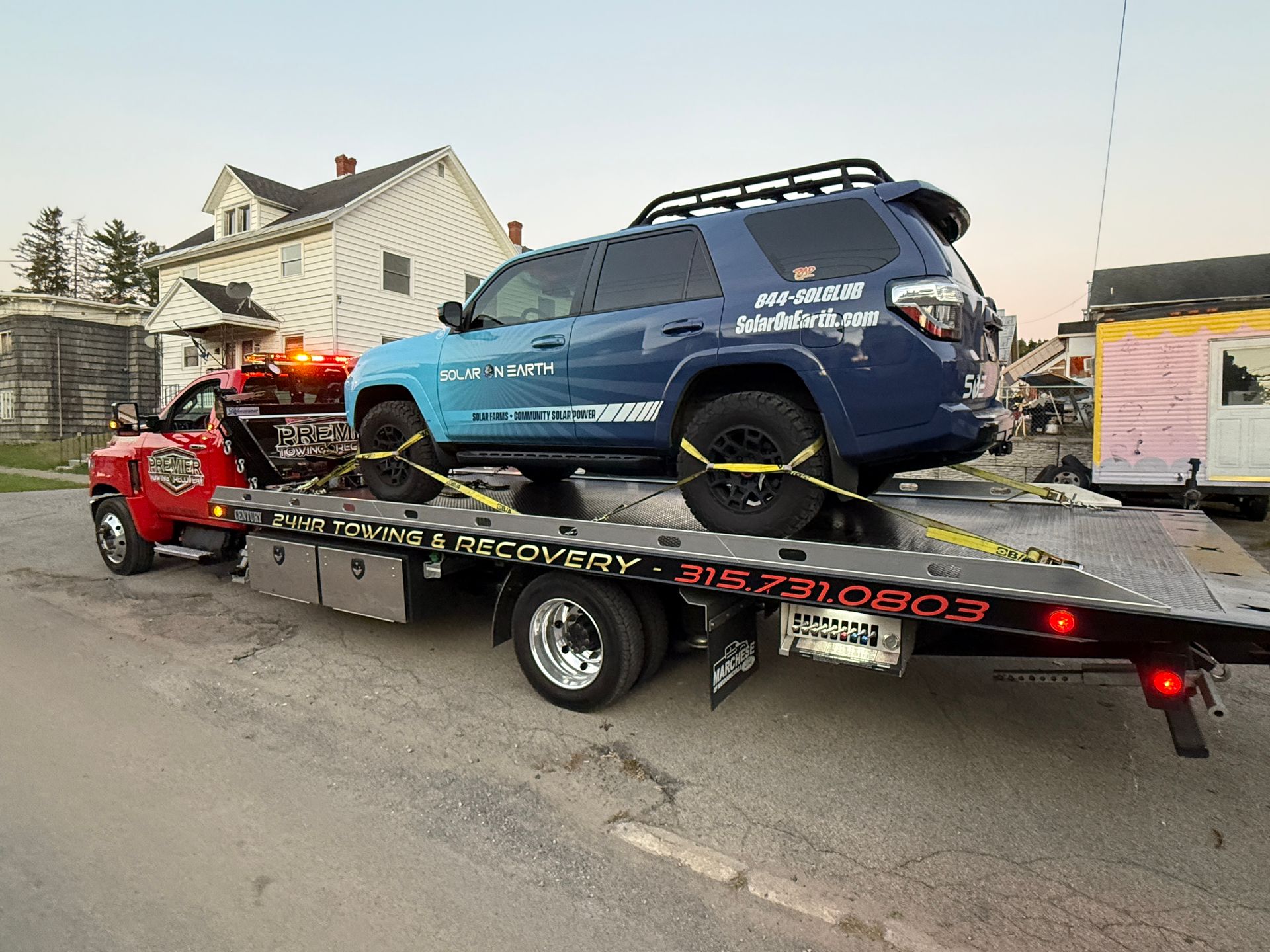 A blue SUV secured on the flatbed of a red tow truck parked on a paved street near a white house.