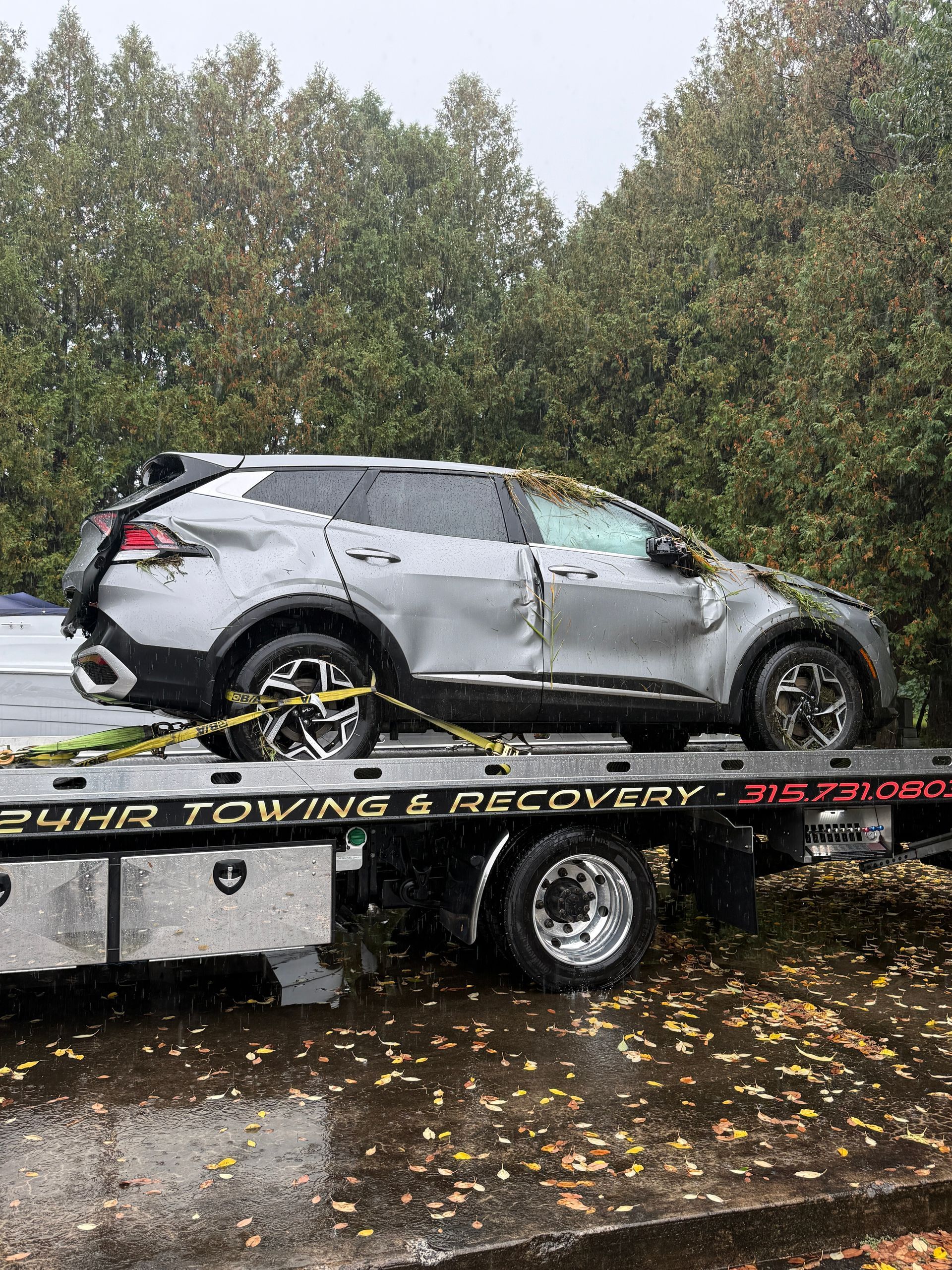 A silver SUV with significant side damage secured on a flatbed tow truck parked on a wet, leaf-covered road.