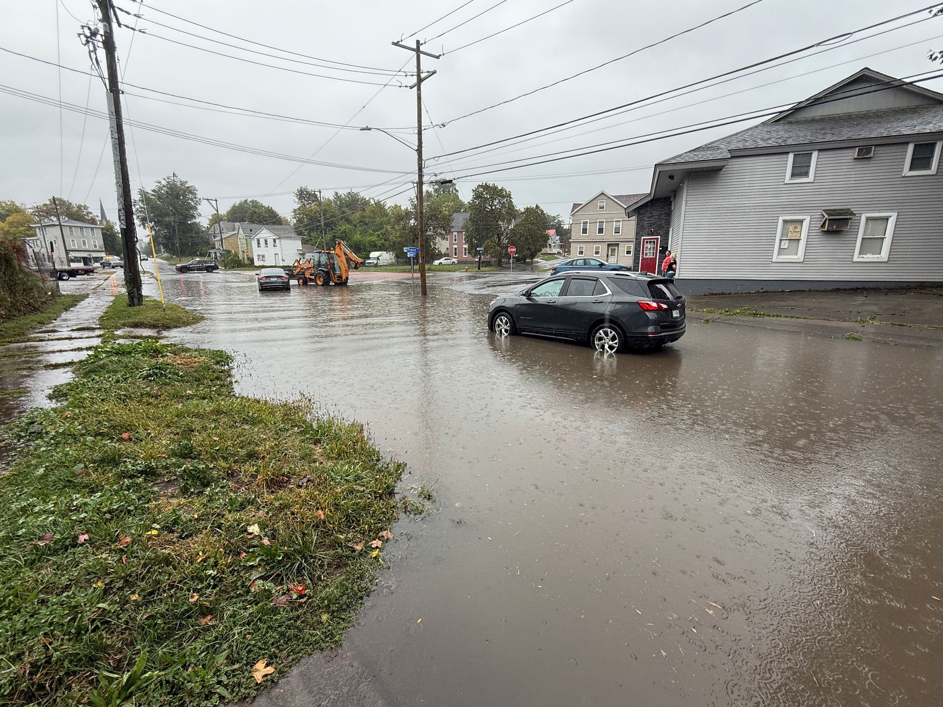 A flooded street with a grey car partially submerged in water, near a house and utility poles on a cloudy day.